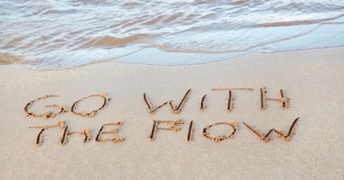 “Go with the flow” written in sand as waves wash toward the shore for “Faith in the Flow”