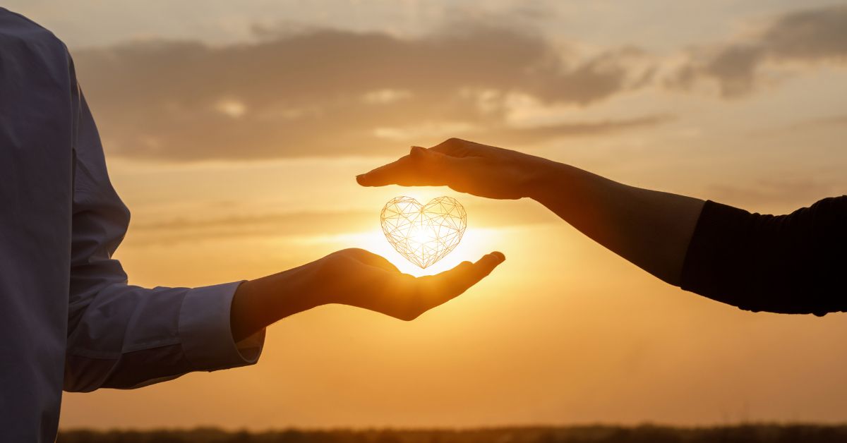 Two people holding a glowing heart shape between their hands at sunset, representing healing through shared care and connection.
