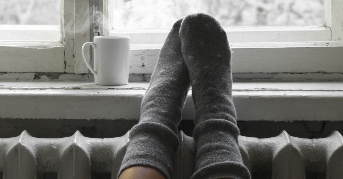 Socked feet resting on a radiator beneath a window, with a steaming mug nearby, capturing a quiet winter moment of warmth and rest.